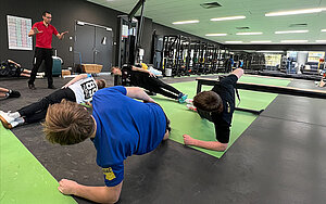 Menschen bei Fitnessübung in einem Trainingsraum, Trainer gibt Anweisungen.